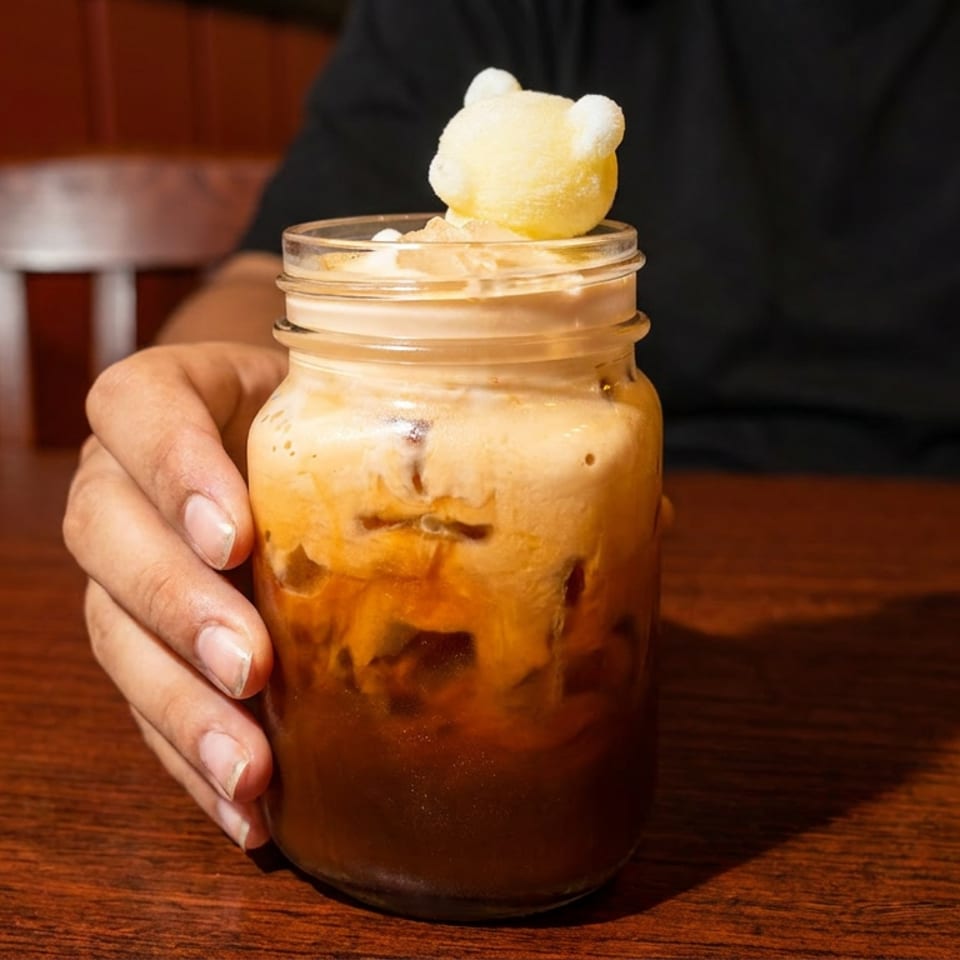 Hand holds a mason jar dessert layered with caramel sauce and cream, topped with a scoop of vanilla ice cream at a wooden restaurant table.